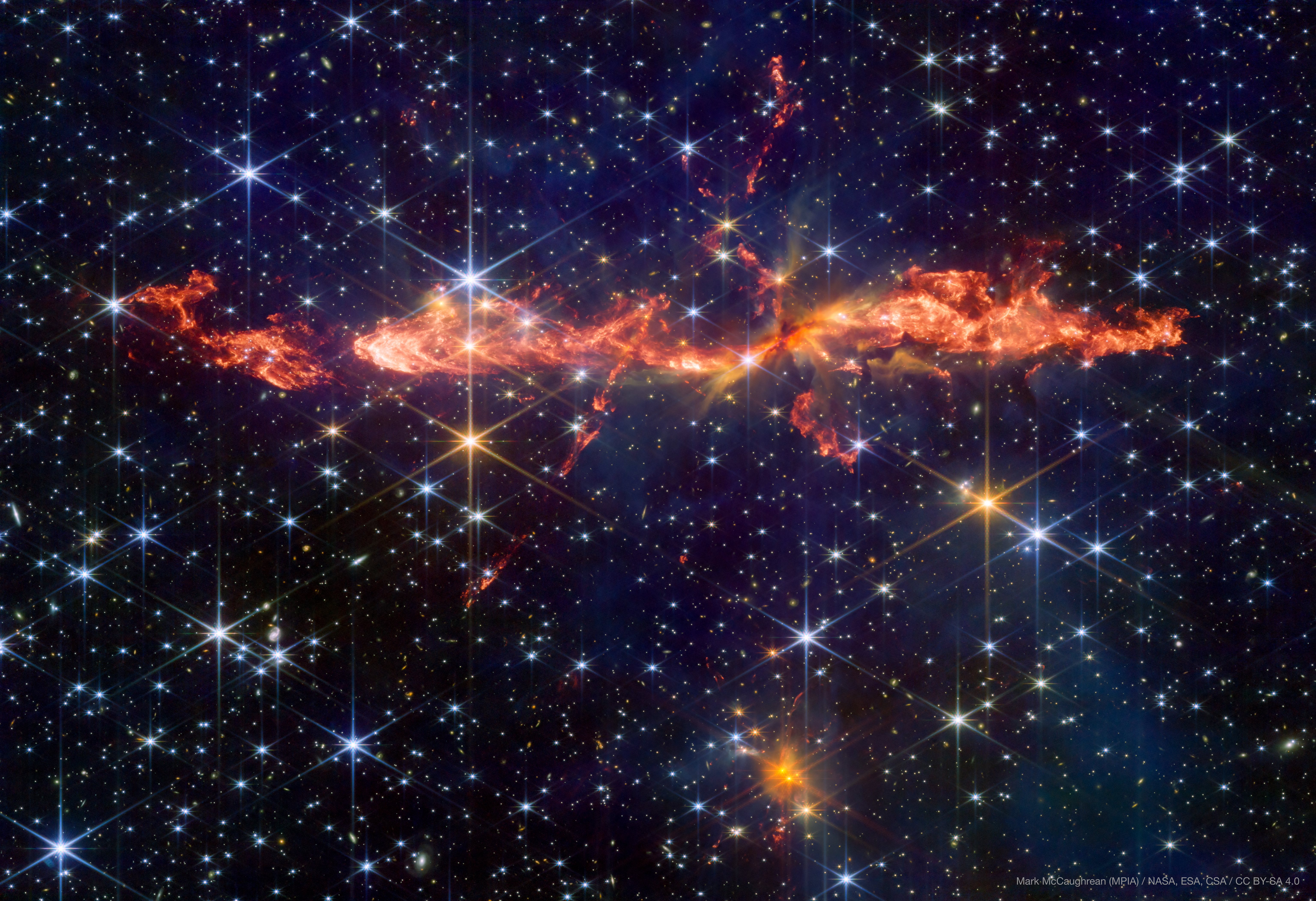 the background of the image is filled with stars. In the center a large, mostly stright jet that looks like flames is visible. smaller jets criss cross this one. At the bottom oa smaller cluster also has outflows. the background of the image is filled with stars. In the center a large, mostly stright jet that looks like flames is visible. smaller jets criss cross this one. At the bottom oa smaller cluster also has outflows.