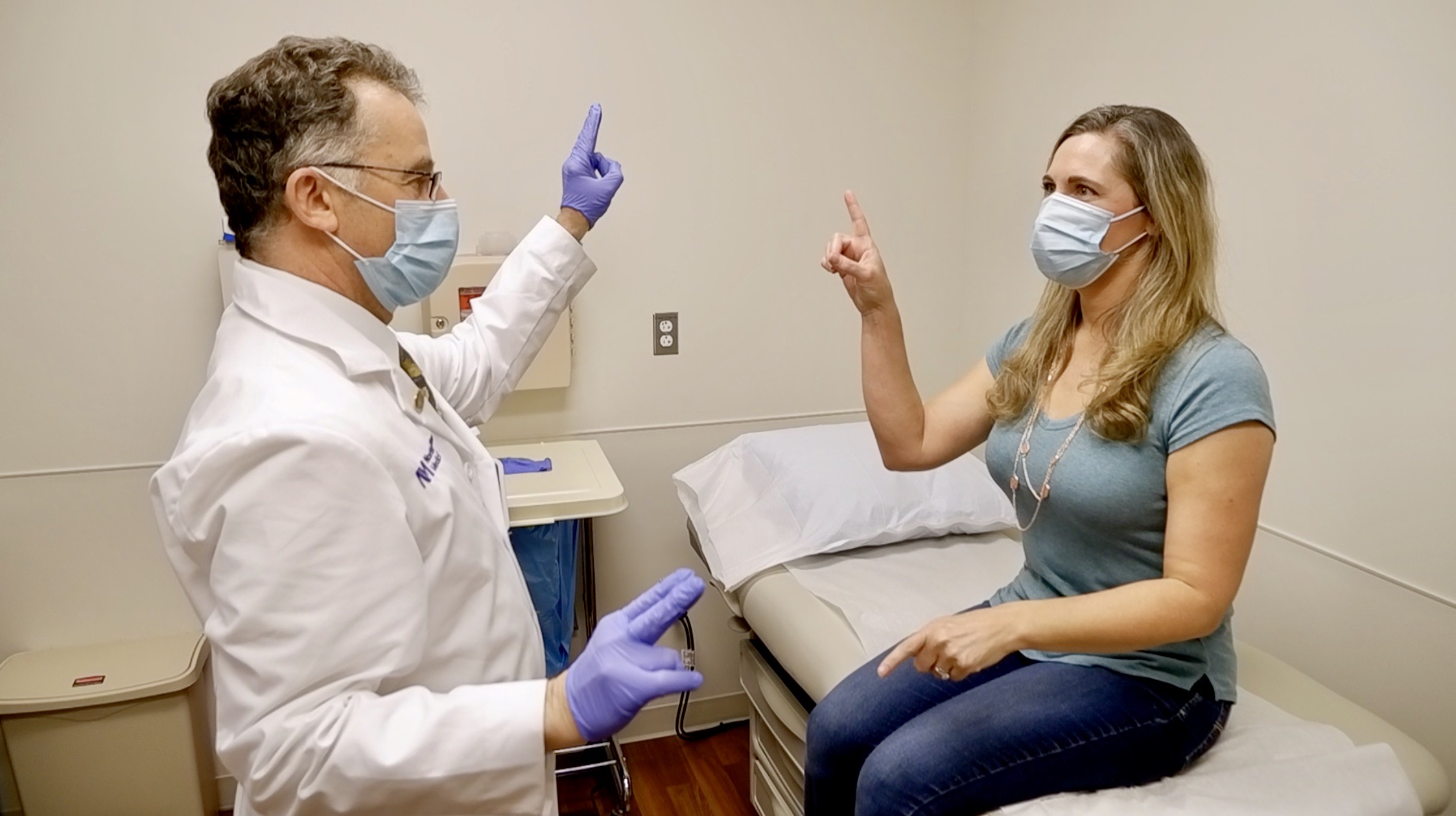 male doctor wearing a white coat, surgical mask and purple gloves meeting a patient with long blond hair and a surgical mask who is sitting on a bed in the examination room