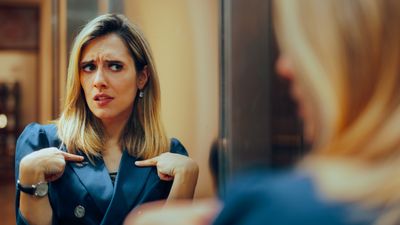 A woman in a blue blazer looks in a mirror pointing at herself with both hands and looking insecure.
