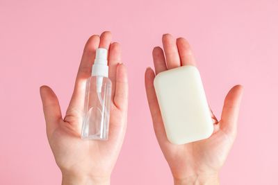 two hands on a pink background; the left is holding a clear spray bottle of hand sanitizer and the right a white bar of soap