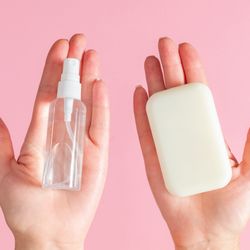 two hands on a pink background; the left is holding a clear spray bottle of hand sanitizer and the right a white bar of soap