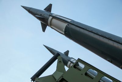 A photo taken under two defense missiles pointing at the sky. The missiles enter the frame from the bottom right and extend to the top left. They are a dark green color with a white band towards their top. 