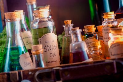 A bunch of glass bottles with colourful liquids on a shelf with handwritten labels that say things like 'Best Ever Snake Oil. No Really!'