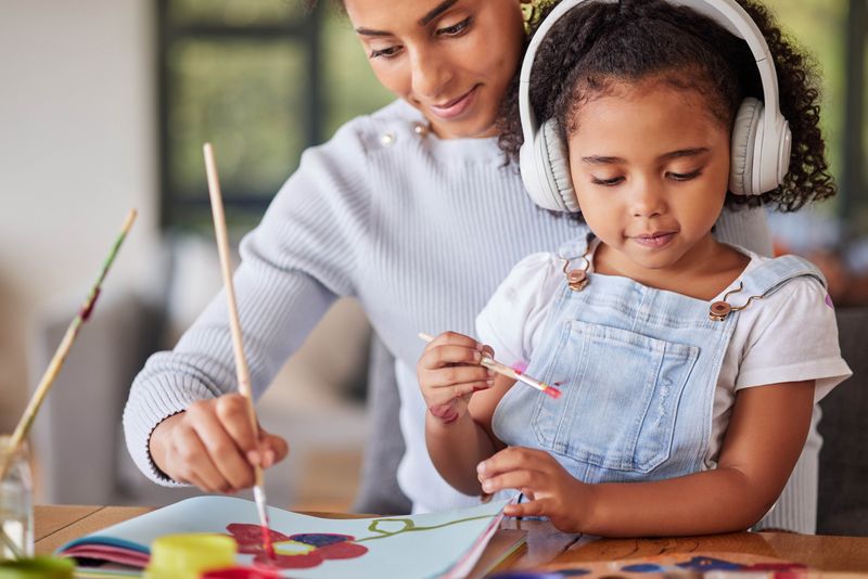 A photo showing a woman and a little girl painting at a table. The little girl is sat on the woman's lap. They both have paint brushes in their hands and are working on a colorful flower painting. The little girl is looking slightly to her left and she's wearing headphones. 