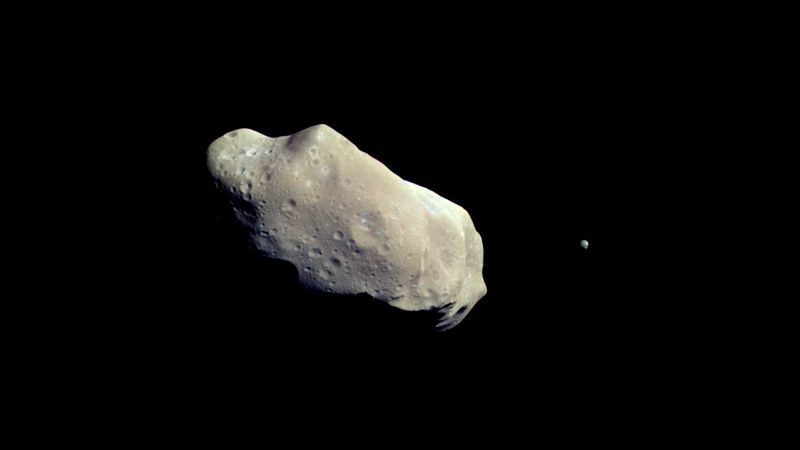 asteroid ida is potato shape and seemingly stony with many craters on its surface. a small rocky dot is around it.