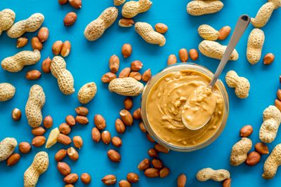 flat lay of jar of crunchy peanut butter on a bright blue table surrounded by scattered peanuts in and out of their shells