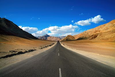 A photo taken of a road passing through some arride yellow hillsides. The road appears to curve downwards slightly before lifting towards the horizon. 