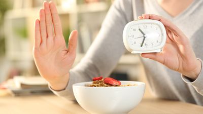 A photograph showing a woman with a bowl of cereal holding up a clock.