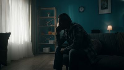 A photo of a middle aged man sat on the edge of a bed in a darkened room. The man's shape is mostly a silhouette and he resting his face on his left hand as if in contemplation. The curtains to the bedroom are drawn and the space is weakly illuminated by two small lamps in the background.