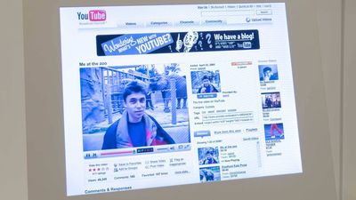 A photo showing a computer screen with the restored 2006 YouTube page with "Me At The Zoo" on it. The video shows a young man standing in front of an elephant enclosure on the grey day. The video is surrounded by suggested videos and also other features such as rating the video.