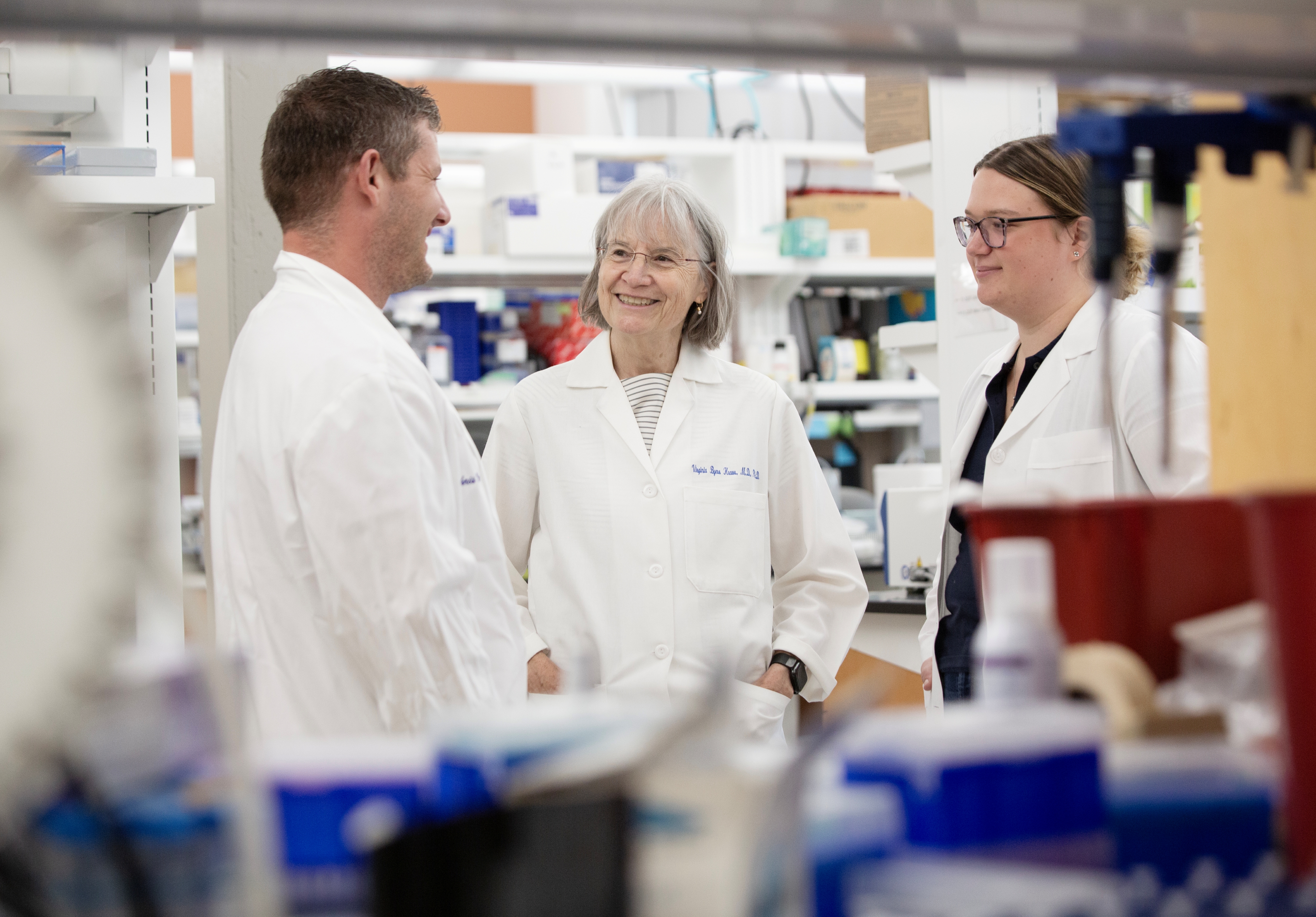 three scientists speaking in a lab, one white man and two white women, both wearing glasses, the older woman in the middle is smiling and wearing a white lab coat