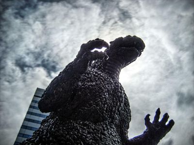 godzilla statue photographed from below with a cloudy sky and a high-rise building in the background