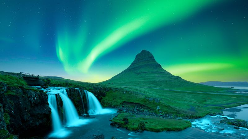 a beautiful waterfall is seen on a green landscape in iceland with a bright green aurorae overhead.