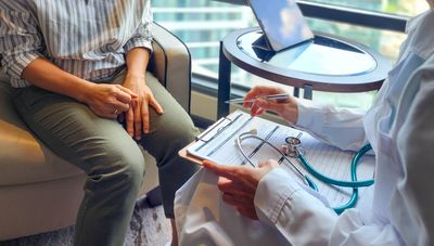 A photo showing a woman sitting opposite a doctor holding a clipboard on her lap. The photo is framed so that only the torsos of the two seated women are visible. The patient is wearing green trousers and has a striped top, while the doctor is wearing a white coat.
