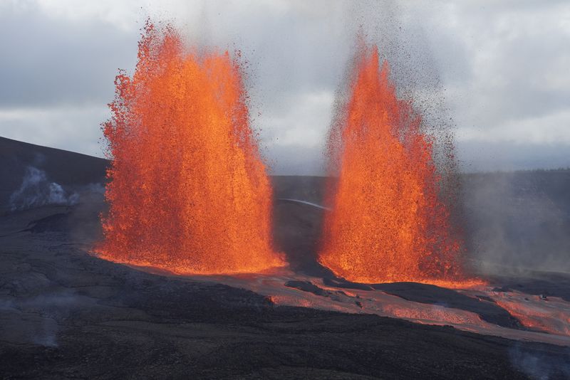 A photo showing two lava fountains on Kilauea summit. They appear as bright orange and red plumes set against a dark brown and black volcanic earth. There are two magma channels flowing to the bottom right of the image, extending from their bases.