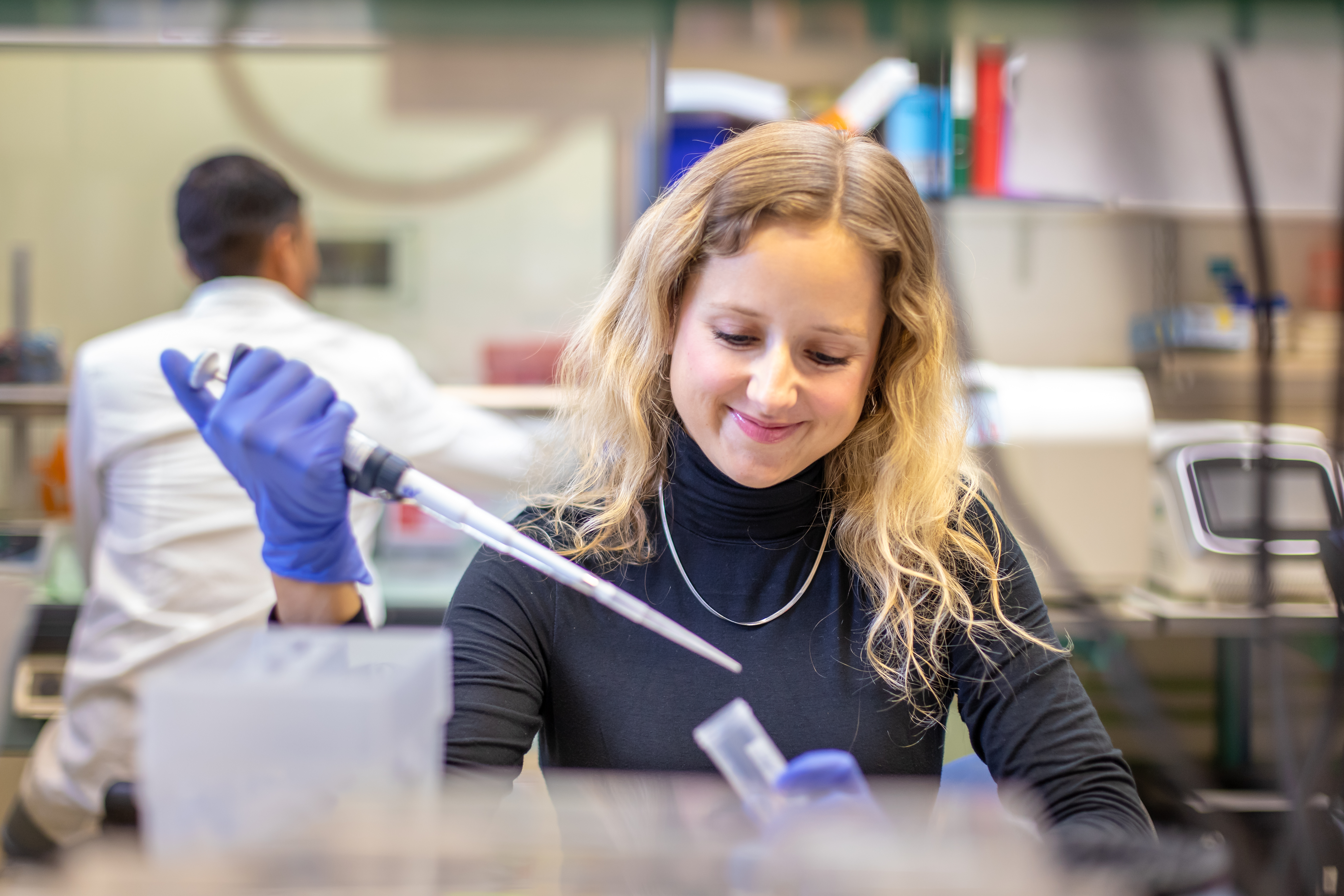 A blonde woman wearing a grey turtleneck sitting at a lab bench using a pipette