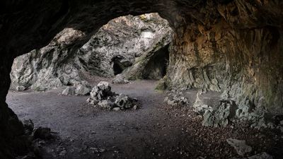 A photo showing a large cave complex with some arches exposed to sunlight. The ground is covered in gravel and stones.