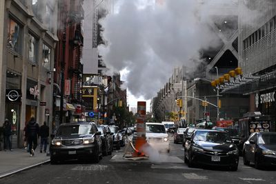 A photo showing a busy street with cars lining up either side of a pipe with plumes of steam floating above it. 