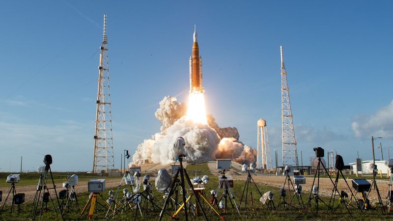 A photograph of Artemis II launching, showing the Space Launch System and Orion Capsule in the initial stages of lift off with media cameras in the foreground.