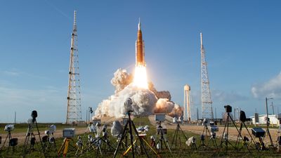A photograph of Artemis II launching, showing the Space Launch System and Orion Capsule in the initial stages of lift off with media cameras in the foreground.