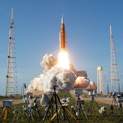 A photograph of Artemis II launching, showing the Space Launch System and Orion Capsule in the initial stages of lift off with media cameras in the foreground.