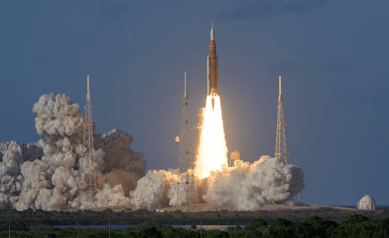 The rocket is see producing fire and vapor as it passes the launch pad tower.