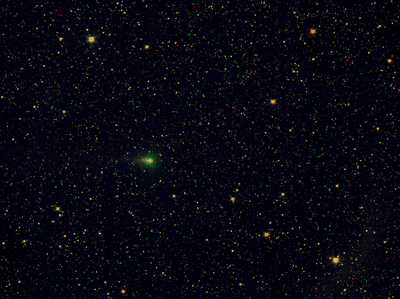 A wide, dark sky is filled with countless tiny points of light, like a dense spray of stars. Near the lower left of centre, a small, soft green glow stands out among the stars, appearing slightly fuzzy compared to the sharp pinpoints around it. This is comet 3I/ATLAS. The rest of the scene fades into deep black space with scattered stars of varying brightness.