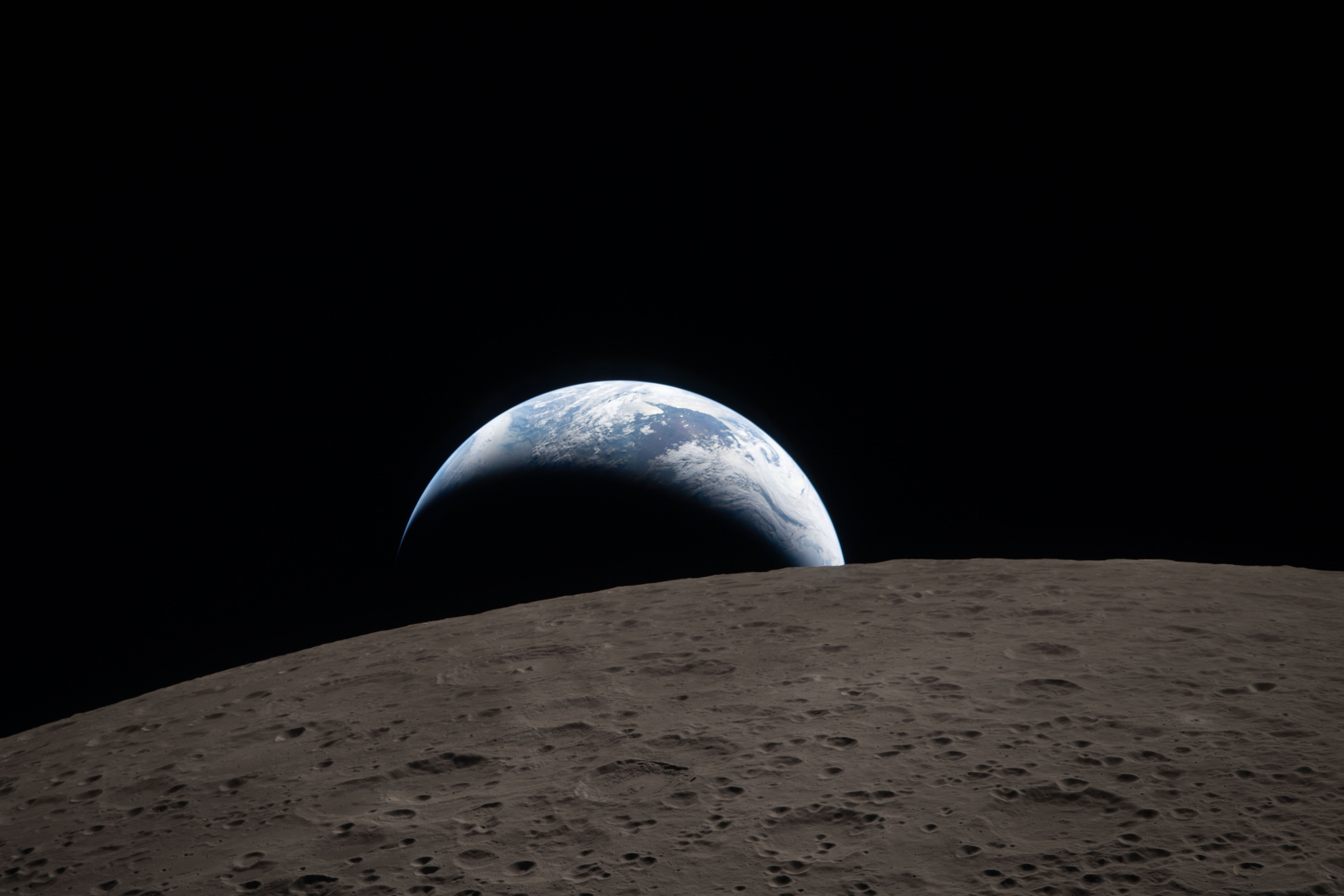 A crecent of the Earth is seen setting behind the crater surface of the Moon