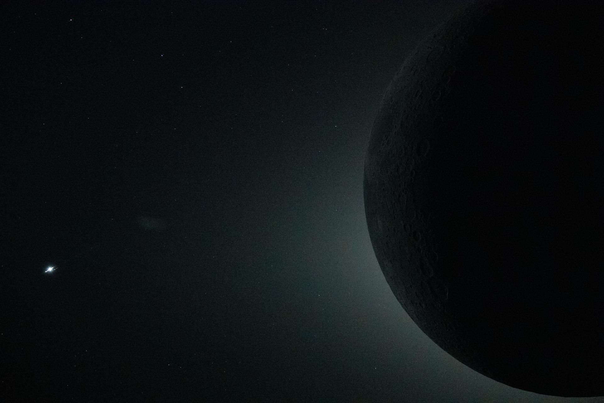 The Moon is mostly dark, some features are seen thanks to earthshine a bright dot is seen in the black sky a fair distance away