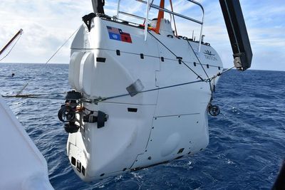 A photo showing a white submersible being held ahead of being put into the water. The vessel is shaped like a suit-case or brief-case and is white, showing distinct panels along its sides. There is a railing visible at the top. 