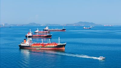 A photo showing several oil and gas vessels anchored in the Strait of Hormuz. They are lined up, fading into the distance on pristine blue water. There is a smaller patrol boat passing the forward most vessel.