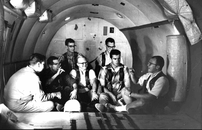 seven men sitting in an empty aircraft, with walls and floor padded.