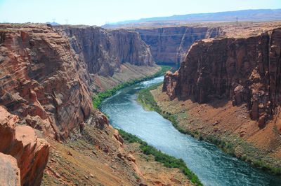A photo taken from the edge of a red-rocked cliff looking down into the canyon with the river running through it. Despite the surrounding terrain being arride and sandy, the banks of the river are verdant. 