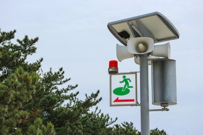 A photo of a tsunami warning system in Japan. The image shows the signal speakers at the top of a pole. There is a sign sticking out of the left hand side showing a person moving in that direction, indicate the evacuation route. 