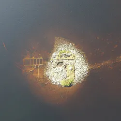 An aerial photo looking down on the small island in the loch. The shot has the near circular island in its center. A small portion of it is above the water, but its larger area is visible in the murky brown waters. A stone causeway can also be seen under the water connecting it to the land to the right of the image.  