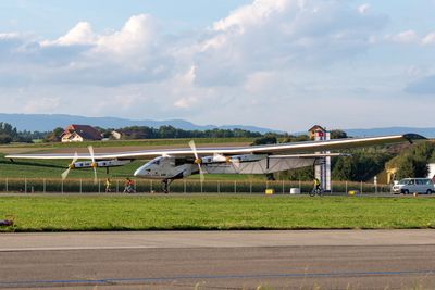 A photo showing the aircraft on a runway in Switzerland. The plane has an angular cockpit and a narrow body that is disproportionately small compared to the large wings that cross from the left to the right of the image. The plane is situated on the edge of a grassy patch, and there are small mountains visible in the background.  