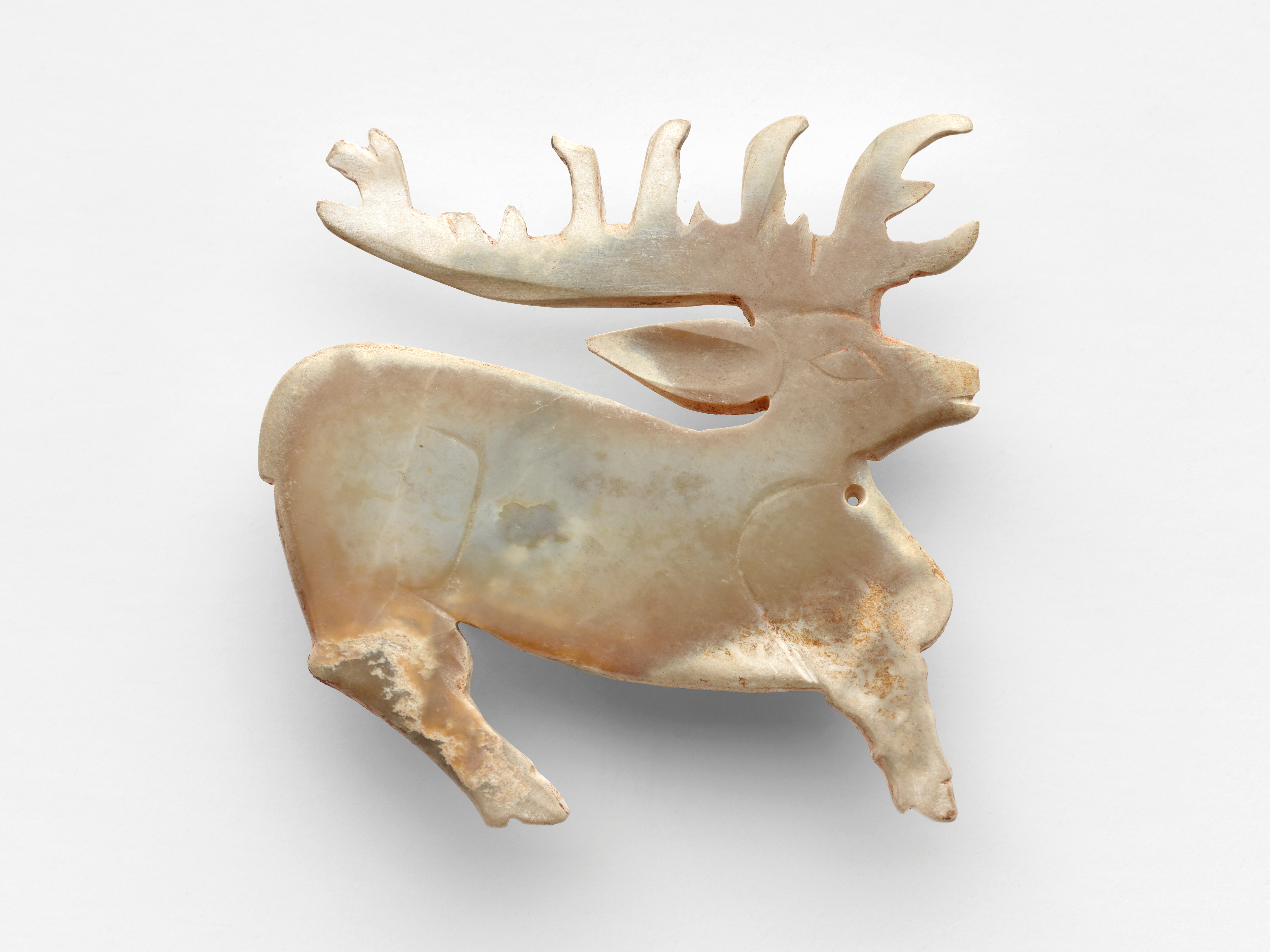 White jade stag presented in silhouette, with its characteristic horns extending backward to repeat the curving form of the stag’s body. Details such as the eye, ear, and haunches are indicated with beveled lines. The animal’s cloven hooves are schematically presented. There is a circular perforation on the stag’s throat. White jade stag presented in silhouette, with its characteristic horns extending backward to repeat the curving form of the stag’s body. Details such as the eye, ear, and haunches are indicated with beveled lines. The animal’s cloven hooves are schematically presented. There is a circular perforation on the stag’s throat.