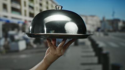 Man holding silver cloche in city street suggests outdoor dining experience with urban backdrop reflecting elegance and service.