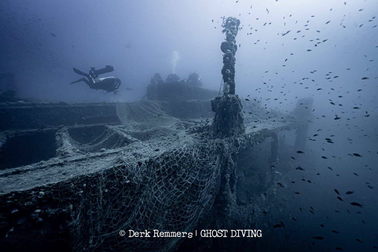 A photo showing a ship wreck at the bottom of the sea. There is a diver swimming just above its surface. The entirety of the ship is covered in tangled fishing nets. A photo showing a ship wreck at the bottom of the sea. There is a diver swimming just above its surface. The entirety of the ship is covered in tangled fishing nets.