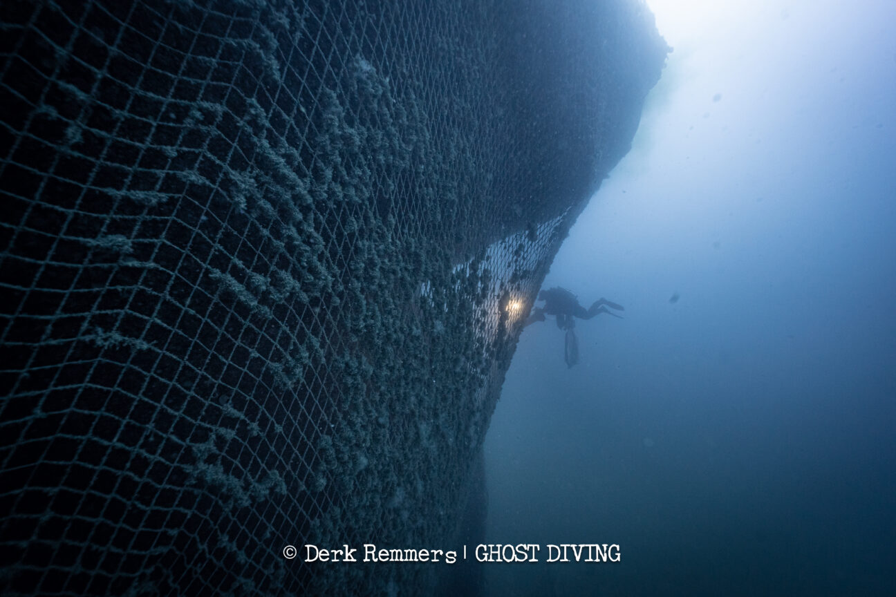 A photo taken from the underside of a shipwreck covered in a large fishing net. The shipwreck takes up two thirds of the shot, revealing just how extensive the abandoned net is. A diver can be seen near the center of the image working on the net that dwarfs them. A photo taken from the underside of a shipwreck covered in a large fishing net. The shipwreck takes up two thirds of the shot, revealing just how extensive the abandoned net is. A diver can be seen near the center of the image working on the net that dwarfs them.