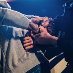 A photo showing a male police officer handcuffing a male suspect from behind. The suspect is wearing a stone grey hoodie. The shot is framed so that neither person's head is visible and it has been taken at night or in the dark. 