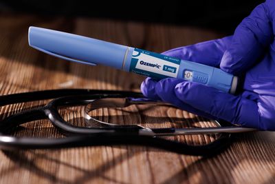 gloved hand holding an ozempic pen over a wooden table with a black stethoscope resting on it