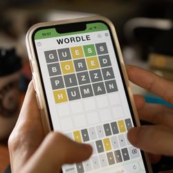 phone screen showing someone playing a game of wordle where they have guessed GUESS, BORED, PIZZA, and HUMAN. There is a coffee cup visible in the background of the shot on a table.