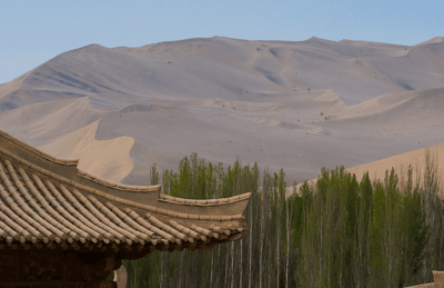The Mingsha Desert peaks over an old-style house and fresh forests in Gansu Province, Northwestern China.