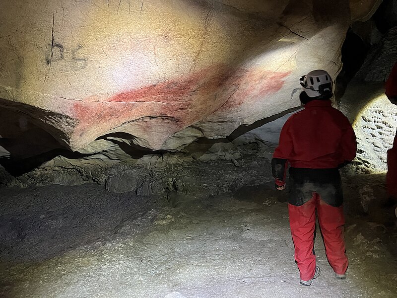 A person in a red jumpsuit and hard hat is looking at a cave wall covered in red ochre. A person in a red jumpsuit and hard hat is looking at a cave wall covered in red ochre.