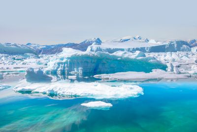 A photo of a sea ice packed around a bay that is melting. The ice has created a green/turquoise hue to the water in the foreground. In the background is an elevated point covered in snow. 