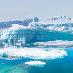 A photo of a sea ice packed around a bay that is melting. The ice has created a green/turquoise hue to the water in the foreground. In the background is an elevated point covered in snow. 