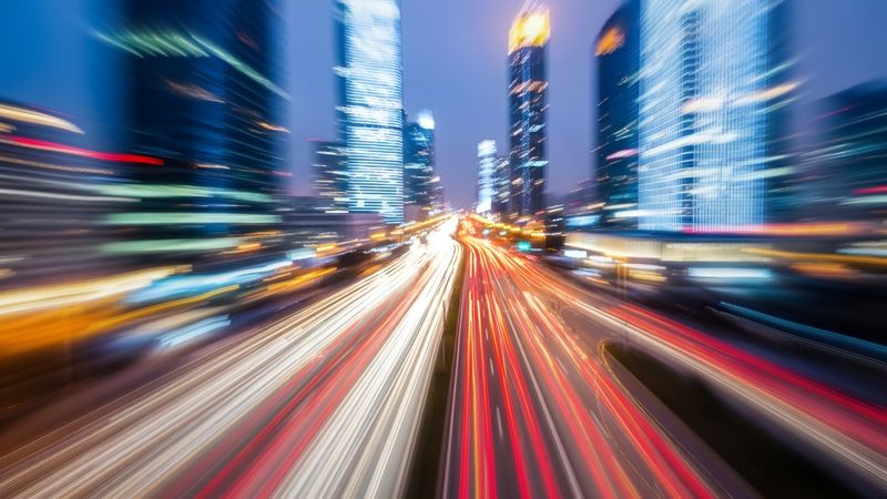 A conceptual image showing a two roads running between some high rise buildings in a city. The exposure has been set so that the lights of the traffic moving along the roads look like streaking lights, giving the image a fast paced, frantic, feeling.