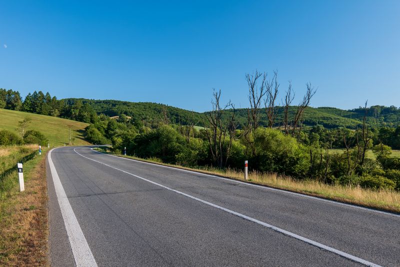 section of road with with line down the middle and at either edge, bordered by grass and bushes with a clear blue sky