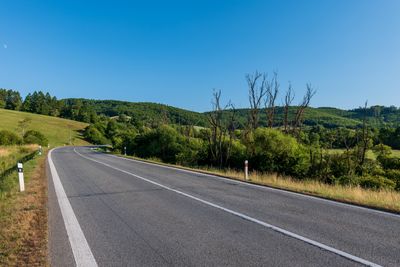 section of road with with line down the middle and at either edge, bordered by grass and bushes with a clear blue sky
